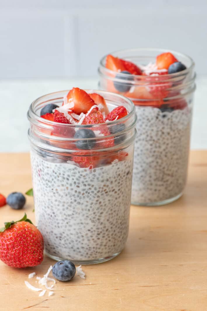 2 jars of chia seed pudding with chopped strawberries, blueberries, and just a few shreds of coconut on top with more fruit around jars.