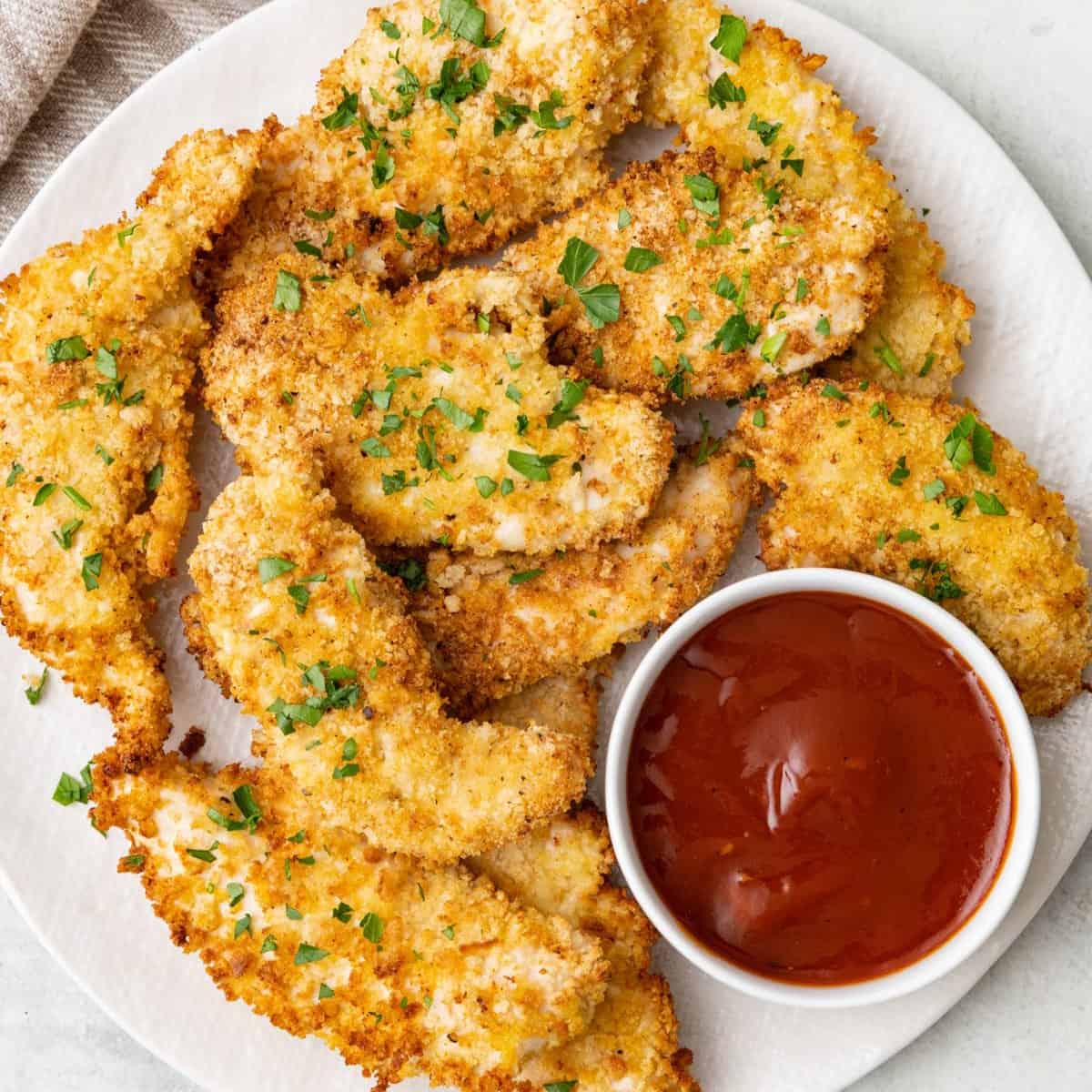 Square image of a plate of crispy air fryer chicken tenders with a small bowl of bbq sauce for dipping.