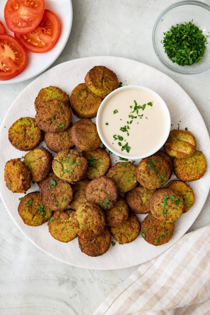 Plate full of air fried falafels with a small dish of tahini sauce and garnished with fresh herbs.