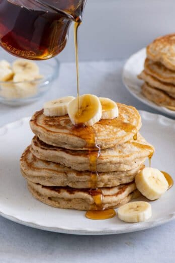 Maple syrup being poured over a stack of banana pancakes with fresh slices of bananas on top.