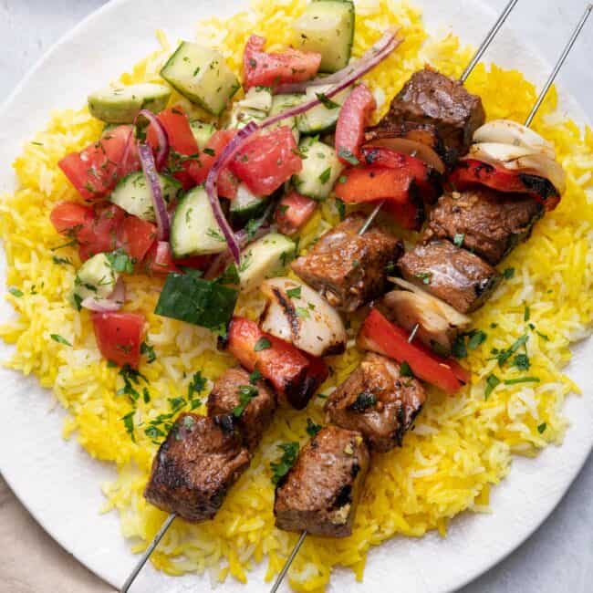 overhead shot of grilled beef kabobs on metal skewers on top of yellow rice and a side of vegetables