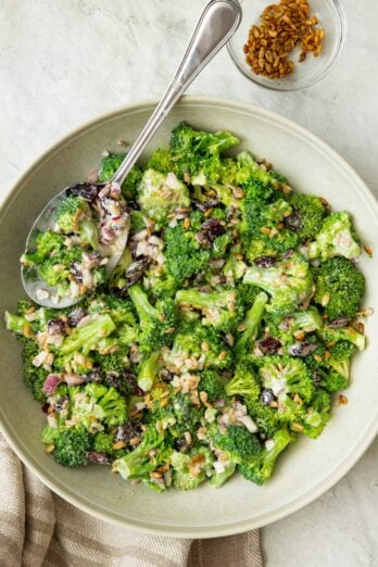 Broccoli salad in a large shallow bow with a serving spoon dipped in and an extra side of toasted sunflower seeds nearby.