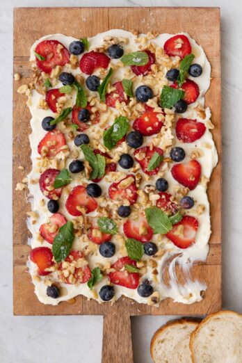 Butter board topped with fruit, nuts, and mit with some smeared off and a few slices of bread nearby.