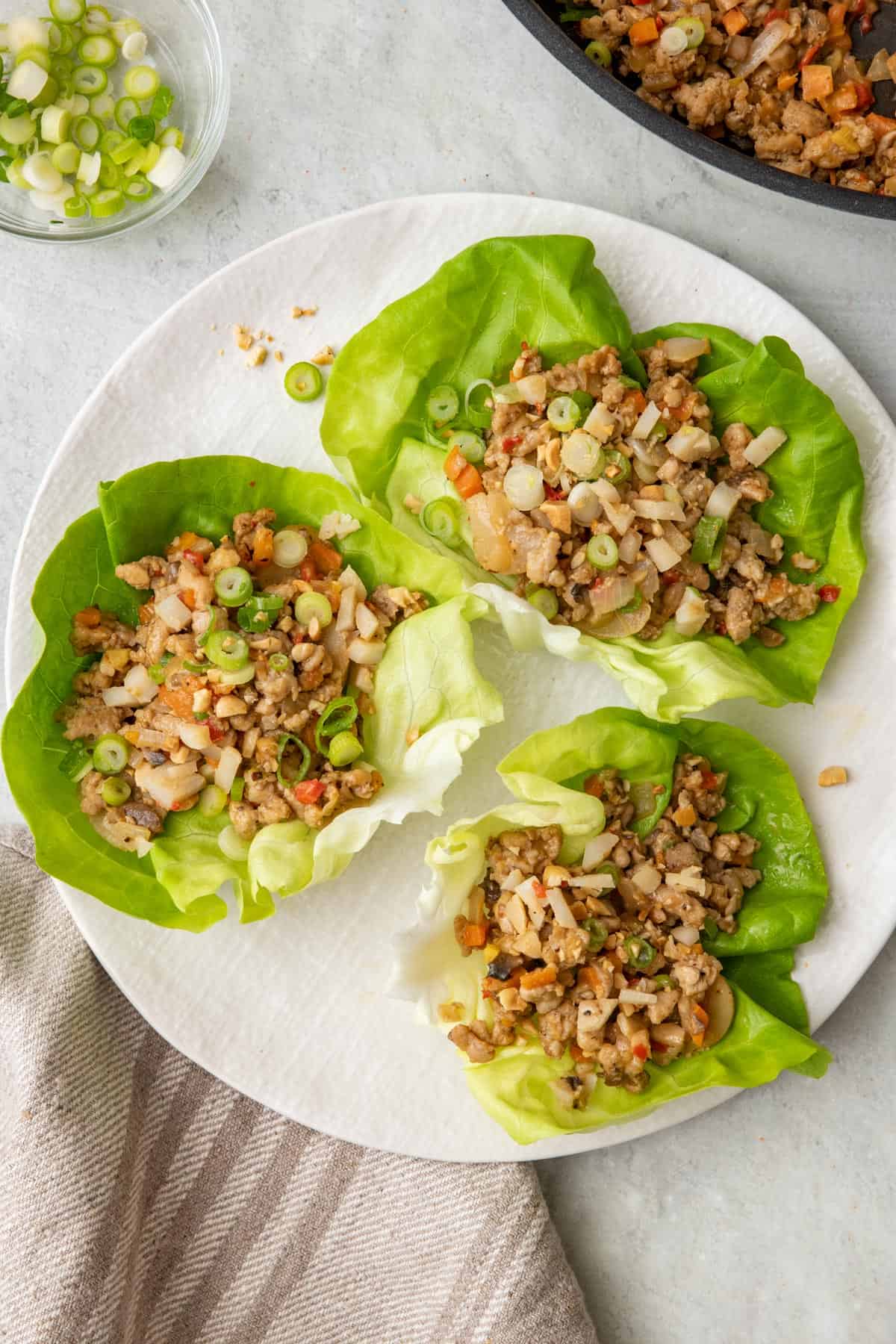 Large plate with 3 butter lettuce and the chicken mixture inside the butter lettuce. On the side are the condiments for serving the chicken lettuce cups including the sauce, peanuts and green onions