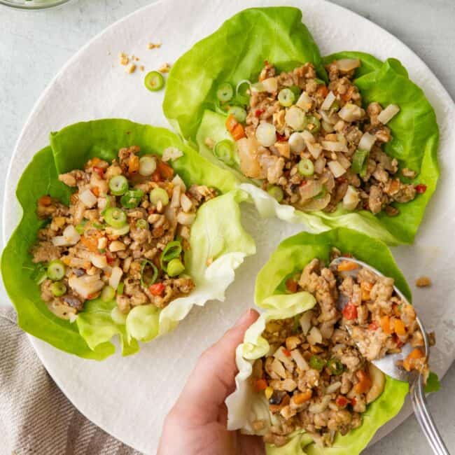 Chicken lettuce cups on a plate with a hand holding one and spooning in the filling to the lettuce cup.