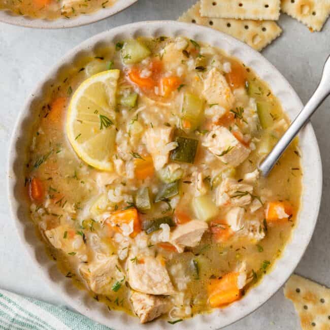 Bowl of chicken vegetable soup garlinished with dill, a spoon dipped inside and crackers nearby.