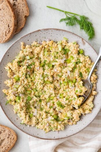 Prepared chickpea "tuna" salad in serving bowl with spoon dipped in, garnished with fresh dill and a few slices of bread nearby.