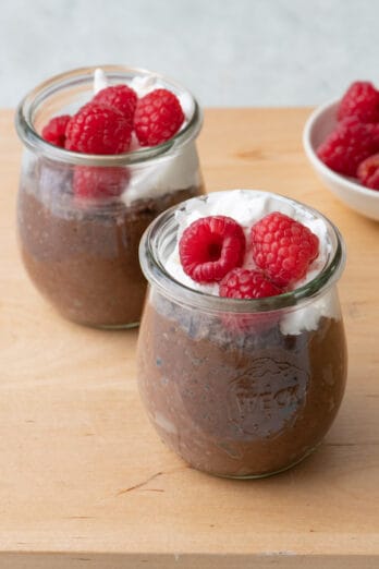 Close up of two glass jars of chocolate pudding topped with whipped coconut cream and raspberries.