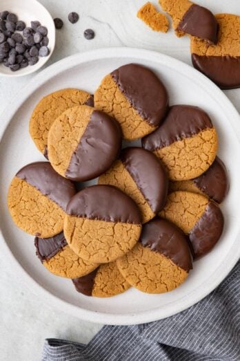 Round plate with cookies stacked on top of eachother with a few off plate and broken and a small dish of chocolate chips.