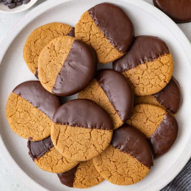 Round plate with cookies stacked on top of eachother with a few off plate and broken and a small dish of chocolate chips.