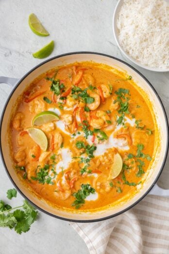 Coconut shrimp curry in pot garnished with a drizzle of coconut milk on top, clinatro, and lime wedges.