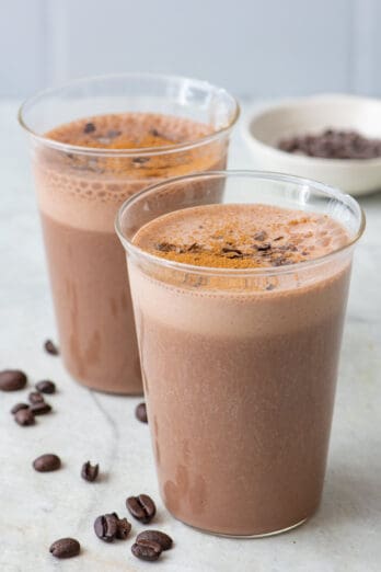 2 small glasses with one in focus both filled with coffee smoothie with a few coffee beans around, garnished with chocolate shavings and a pinch of cinnamon sprinkled on top