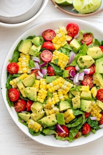 Corn tomato avocado salad in a large white bowl with squeezed limes on the side