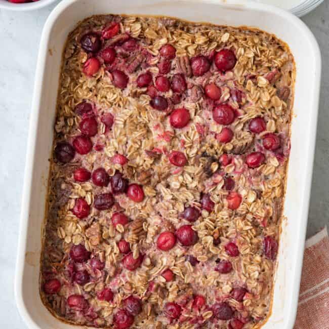 Baked cranberry pecan oatmel in baking dish with fresh cranberries and yogurt nearby in small bowls.