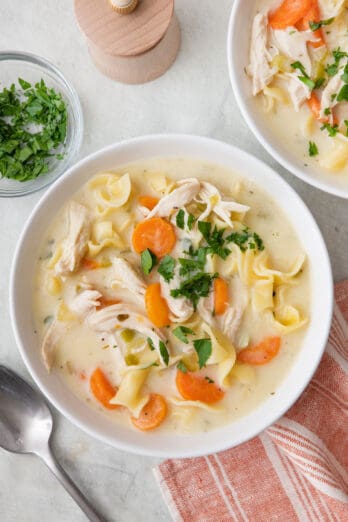 One bowl of soup garnished with pasley with another bowl, a spoon, dish of parsley, and pepper grinder nearby.