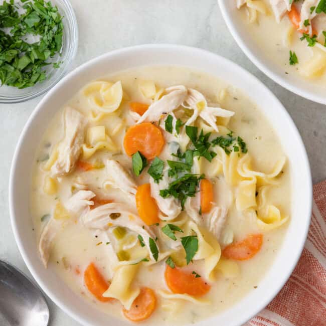 One bowl of soup garnished with pasley with another bowl, a spoon, dish of parsley, and pepper grinder nearby.