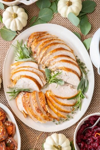 Overhead shot of sliced turkey breast on a large platter with fresh herbs with sweet potatoes and cranberry sauce nearby.