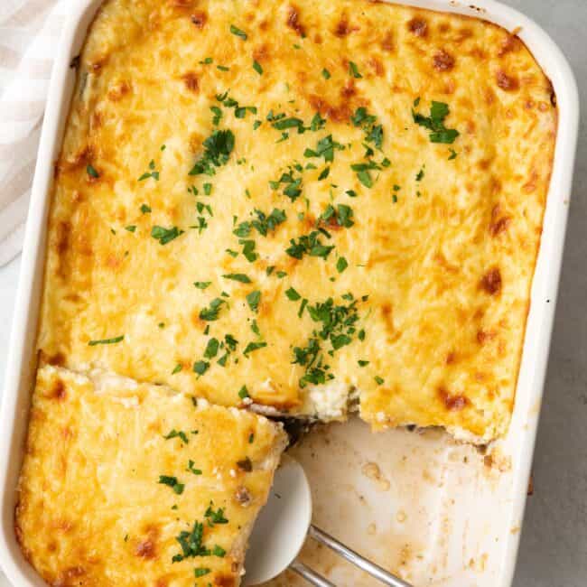 Baked eggplant moussaka with a serving removed on a plate nearby and a spatula lifting up another serving.