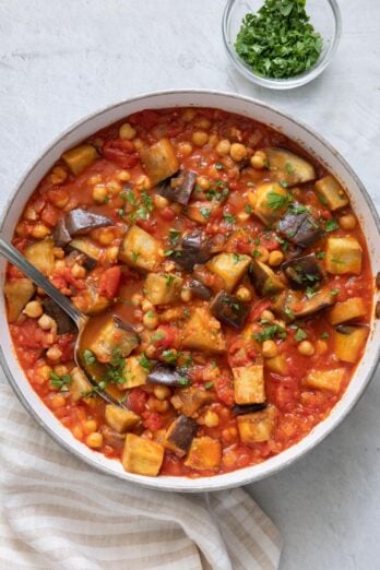 Stew with chickpeas, tomatoes, and eggplant in pot garnished with parsley with serving spoon dipped inside pot.