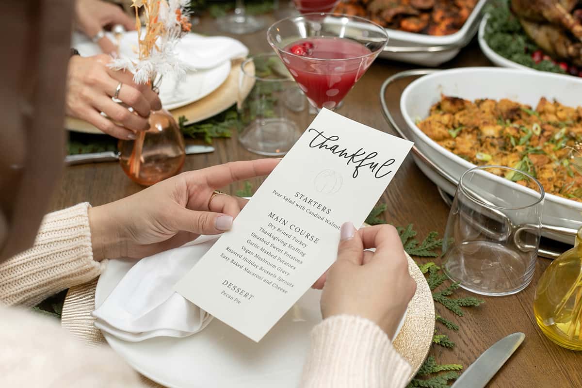 Guest holding Thanksgiving menu listing starters, main coures, and dessert over a dinner plate and table full of prepared recipes from the menu.