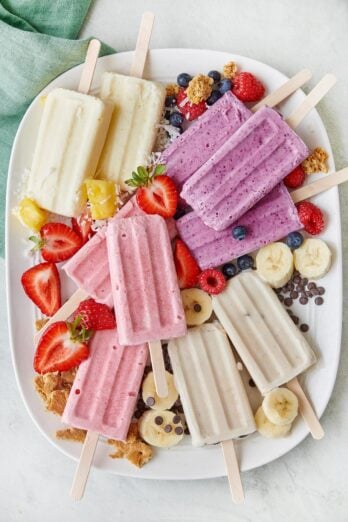 Frozen cottage cheese ice cream popsicles on a large tray: strawberry cheesecake, banoffee pie, pineapple coconut, and mixed berry breakfast pops; extra berries, granola, chocolate chips, and shredded coconut around.