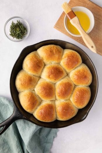 Garlic herb Skillet Dinner Rolls basted with butter in a cast iron skillet with fresh sprigs of rosemary and extra butter and brush nearby.