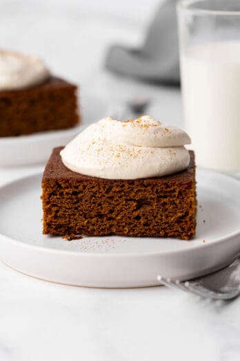 Gingerbread slice on a small plate with brown sugar whipped cream on and cinnamon on top and a small glass of milk nearby.