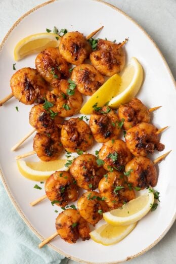 Grilled BBQ shrimp skewers on a large oval platter garnished with chopped fresh parsley and lemon wedges.