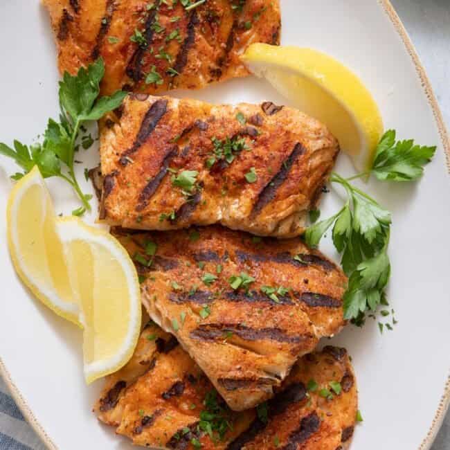 Plater of 4 grilled cod filets garnished with lemon wedges and chopped parsley.