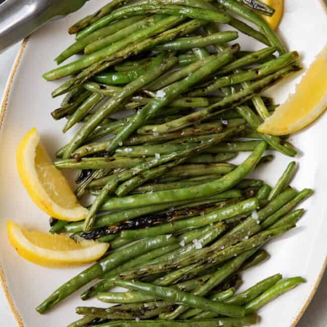 Large platter of grilled greens beans garnished with fresh lemon wedges.