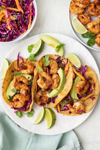 3 grilled shrimp tacos on a plate with sliced avocados, fresh slaw, and lime wedges with a small place of extra grilled shrimp skewers nearby.