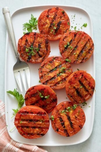 Large white rectangle plate with halved grilled tomatoes and serving fork, garnished with fresh chopped parsley.