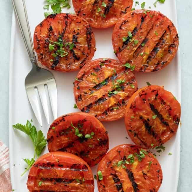 Large white rectangle plate with halved grilled tomatoes and serving fork, garnished with fresh chopped parsley.