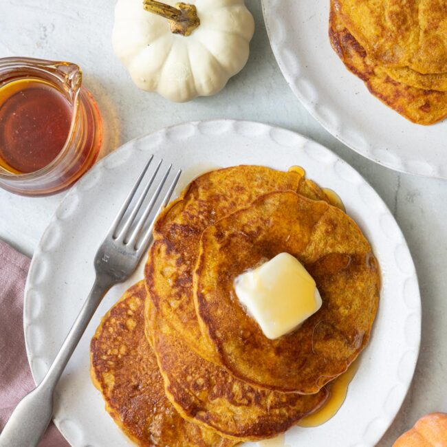 Final plating of recipe on a round white plate with butter and syrup on top, a fork to the side, and small pumpkin decoration around.