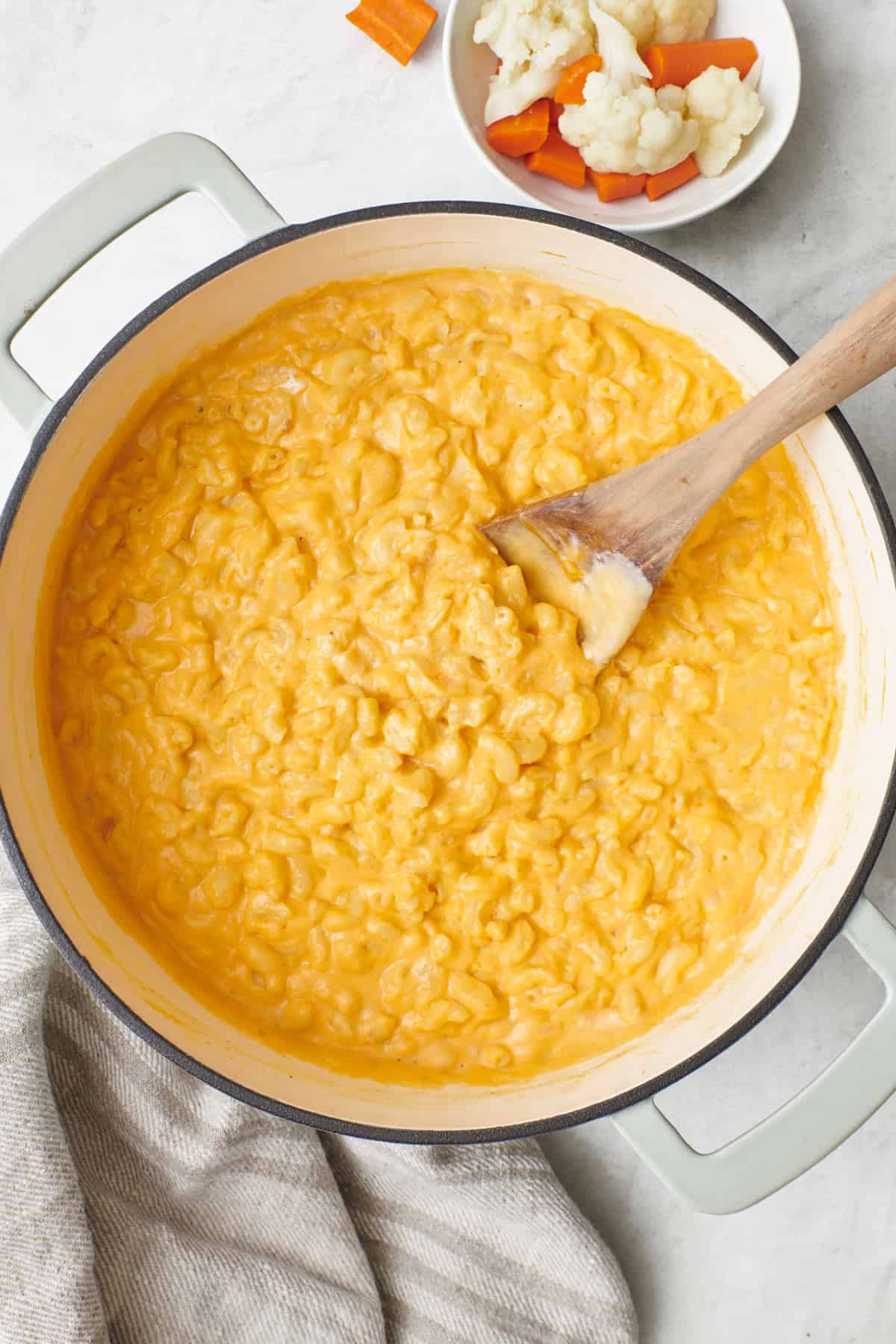 Hidden veggies macaroni and cheese in pot with a wooden spoon dipped in and a small bowl of extra veggies on the side.