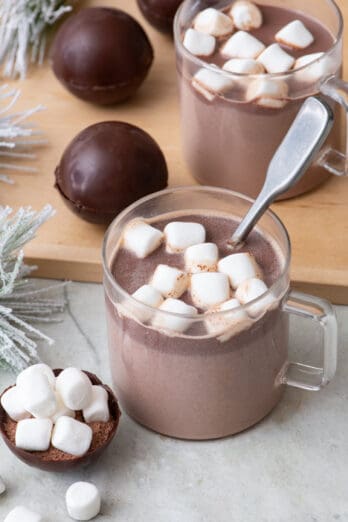 Two glass mugs filled with hot cocoa topped with marshmallows and a spoon dipped in one. Hot cocoa bombs are nearby on a cutting board and one open up front to show inside mix.