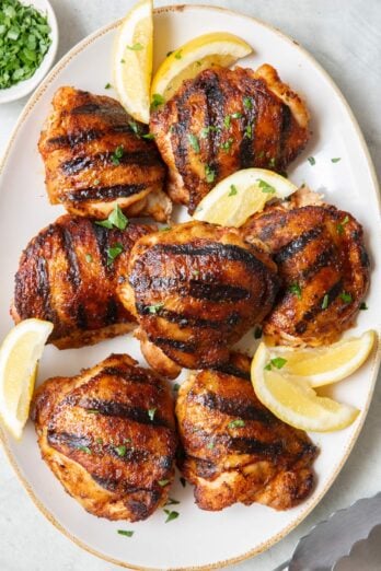 Grilled chicken thighs on an oval platter with crispy skin garnished with fresh parsley and lemon wedges.