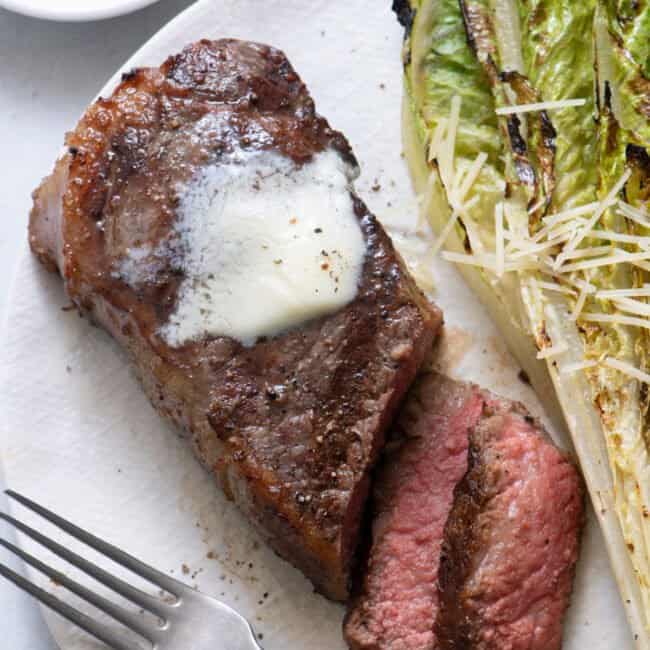 Close up of grilled steak with two slices cut from it topped with melted butter on a large white plate with grilled romaine lettuce, parmesan, and fork.