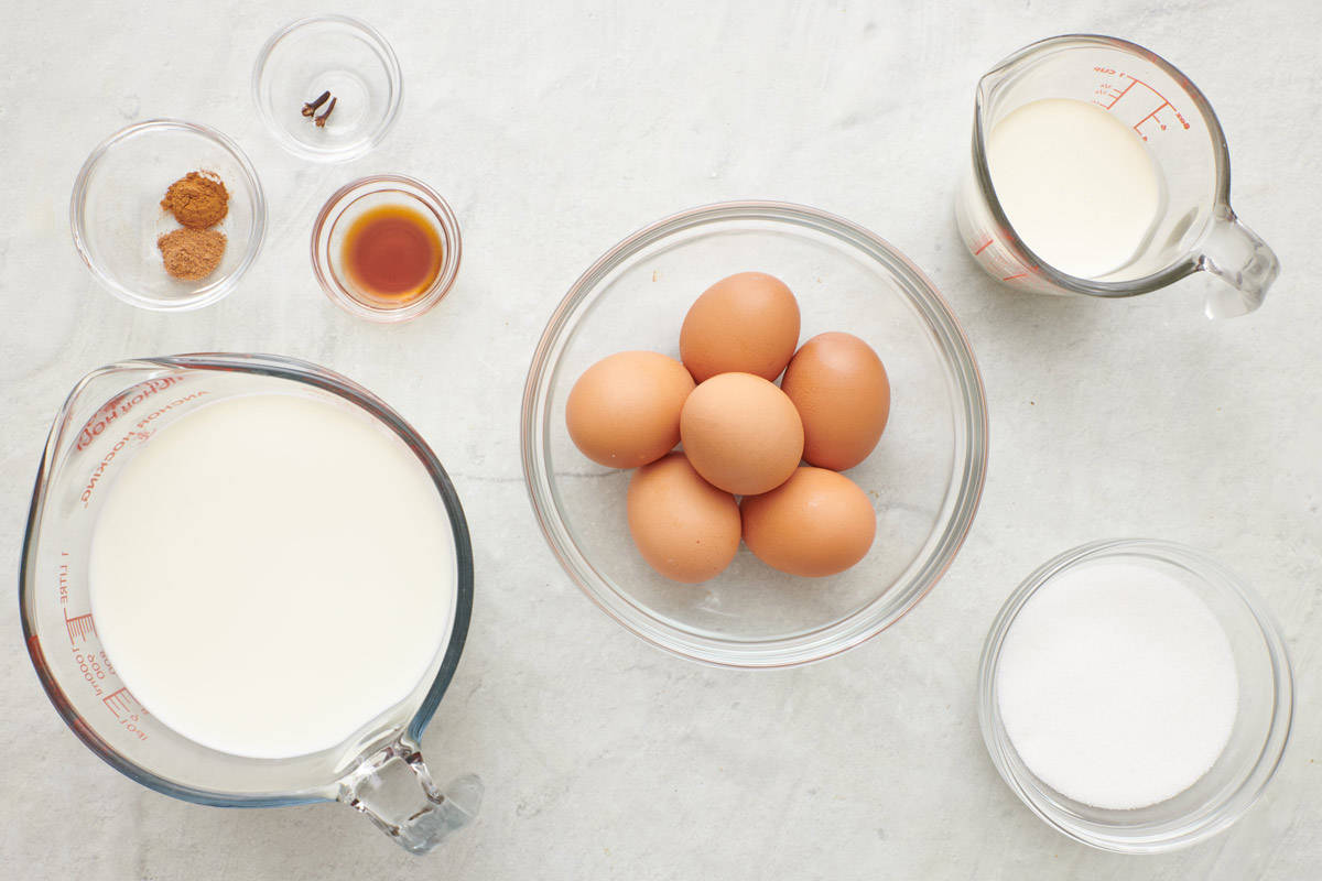 Ingredients for recipe in individual bowls and cups: spices, cloves, vanilla, milk, eggs, cream, and sugar.