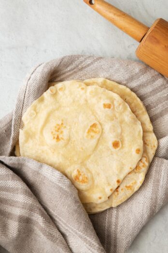 Stack of homemade flour tortillas slightly wrapped in a towel with rolling pin nearby.