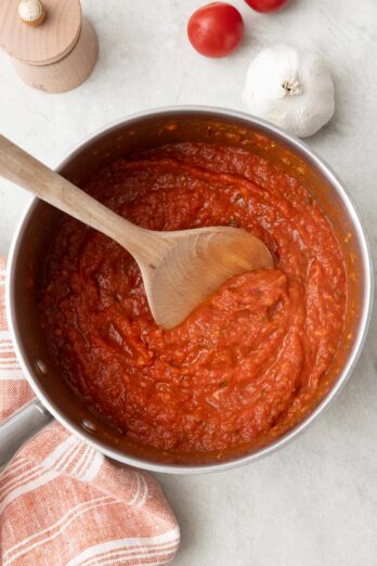 Homemade pizza sauce in a pot with wooden spoon dipped inside, bulb of garlic and whole tomatoes nearby.