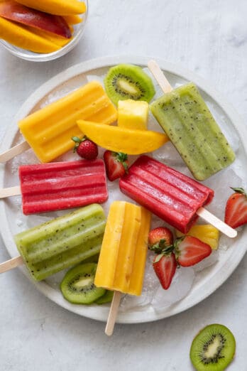 Homemade Mango, Kiwi, and Strawberry popsicles on a large round platter with ice cubes and fresh cut fruit.