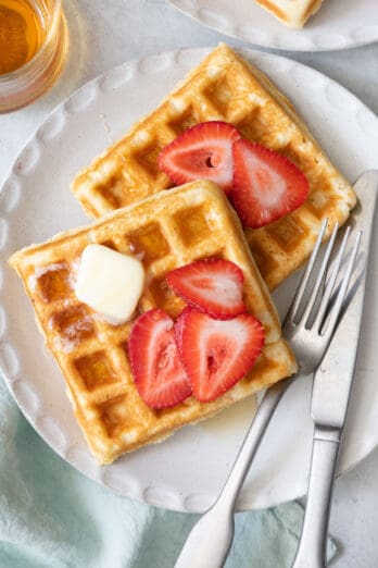 2 square waffles on a white plate with fork and knife, topped with sliced strawberries and butter.