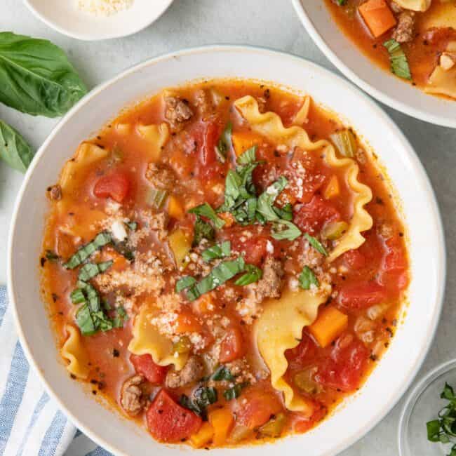 Bowl of lasagna soup garnished with torn basil and parmesan cheese with another bowl nearby.