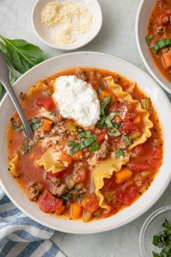 Bowl of lasagne soup with a scoop of ricotta and spoon dipped inside, garnished with torn basil and parmesan cheese.