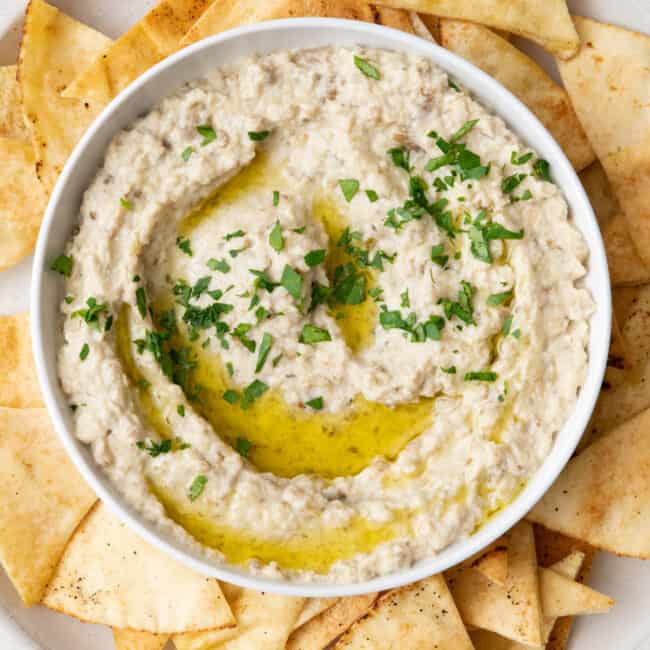 Bowl of baba ghanoush garnished with chopped parsley and olive oil on a platter with pita chips.