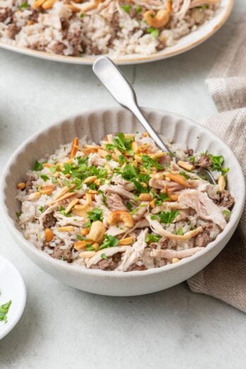 Bowl of Lebanese shreaded chicken and rice with a spoon dipped in and the serving platter nearby. The rice is garnished with toasted nuts and fresh parsley.