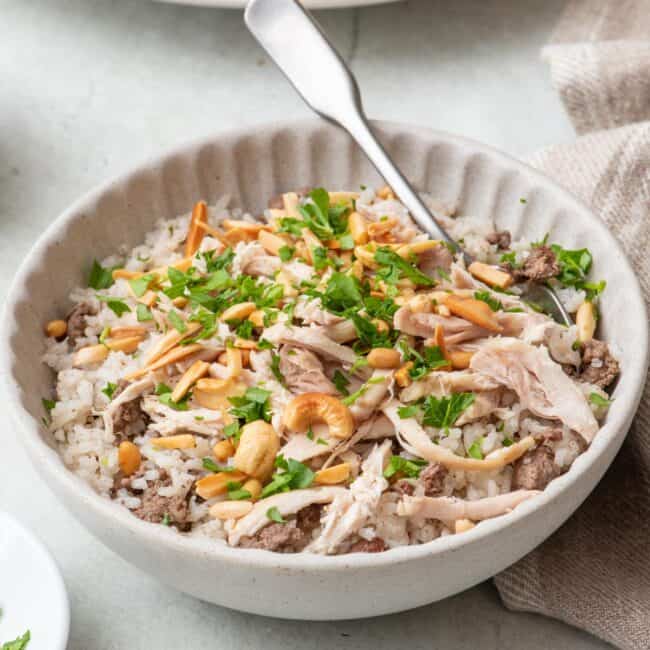 Bowl of Lebanese shreaded chicken and rice with a spoon dipped in and the serving platter nearby. The rice is garnished with toasted nuts and fresh parsley.