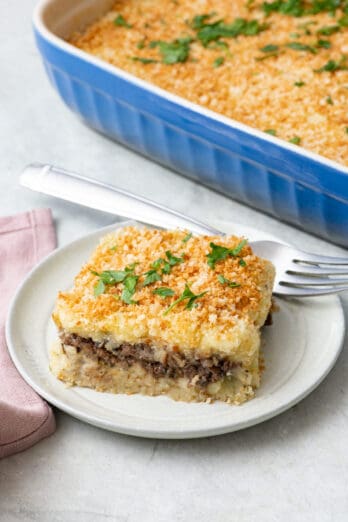 A serving of potato souffle on a small plate to show a top and bottom layer and spiced beef layer in the middle. Baking dish nearby.