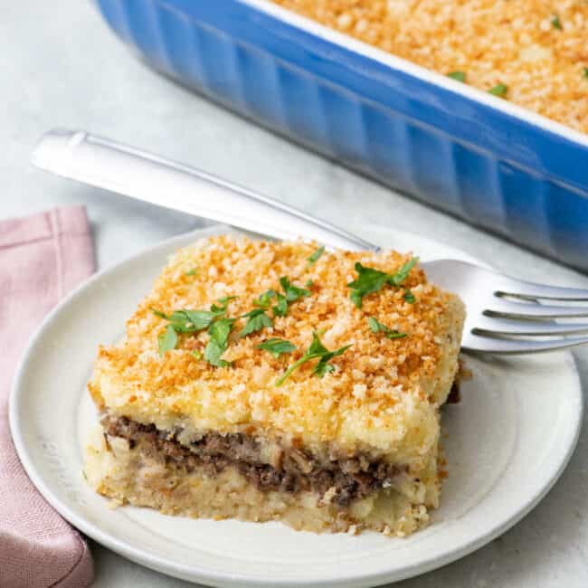 A serving of potato souffle on a small plate to show a top and bottom layer and spiced beef layer in the middle. Baking dish nearby.